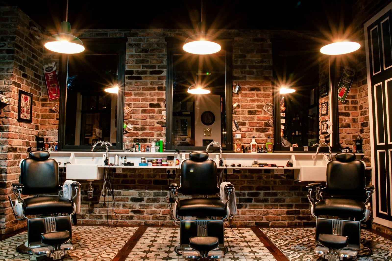 Intérieur du salon : fauteuils de barbier en cuir noir, murs en briques et suspensions dorées